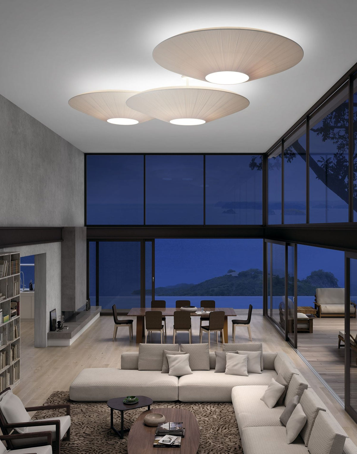 Three Clérachic pleated ceiling lights in ivory at different heights above a lounge with a cream sectional, a dining table and a night-time sea view.