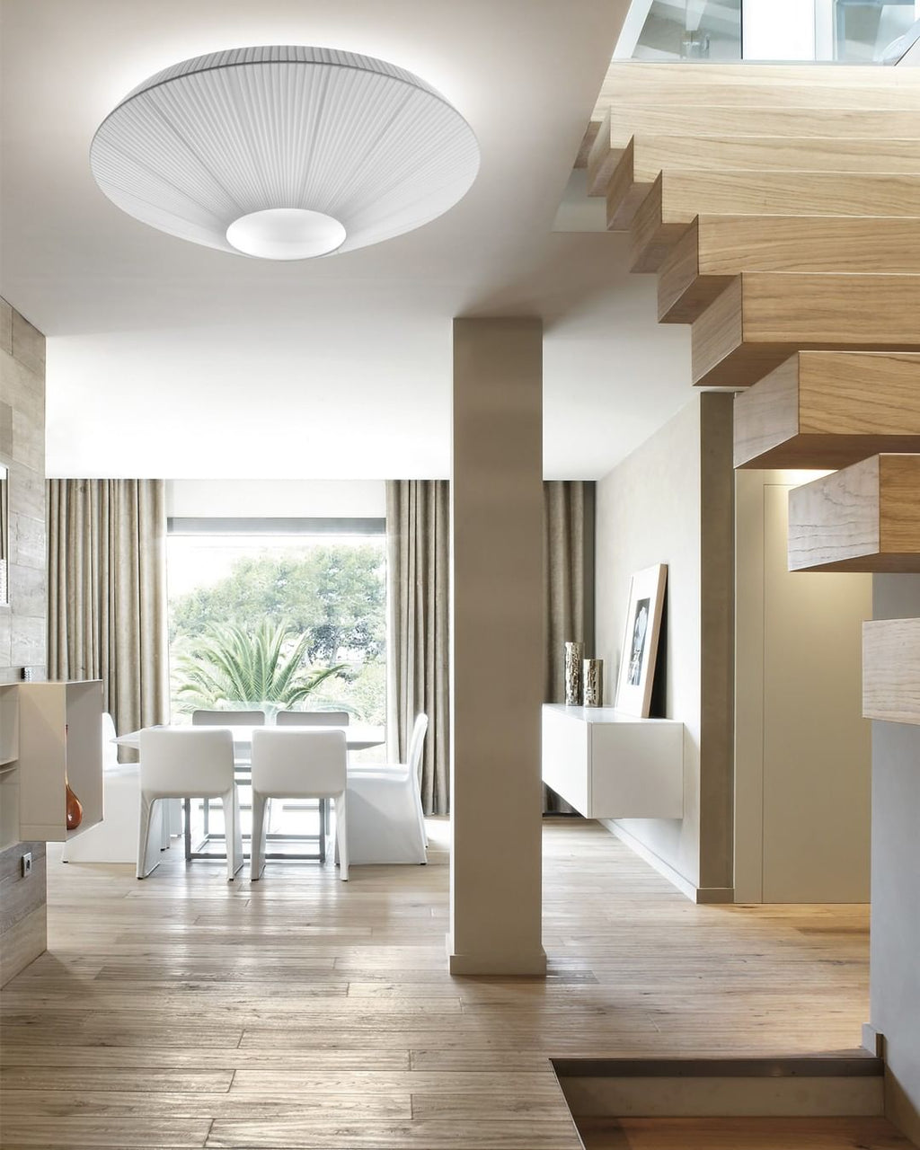 White Clérachic pleated ceiling light above a staircase with floating wooden treads, with a dining table by a window beyond.