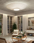 A Clérachic pleated ceiling light in ivory above a living room with French doors, a wooden lounge chair, a coffee table and a tall plant.