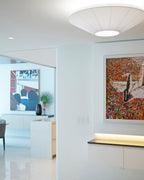 White Clérachic pleated ceiling light in a bright white room with a large colourful painting and a lit shelf below.