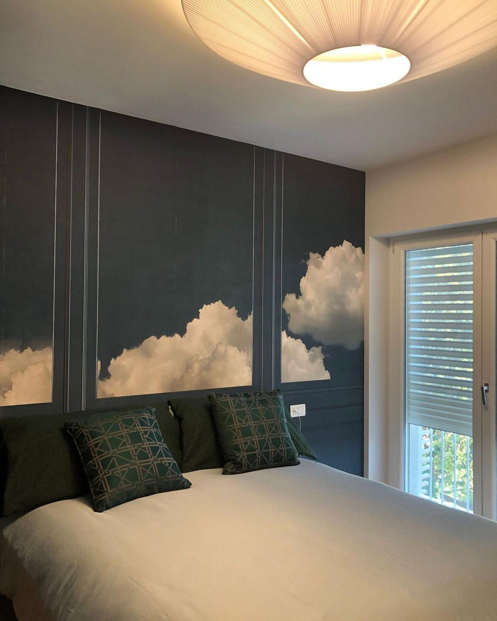 Underside of a Clérachic pleated ceiling light above a bed with dark cushions, in front of a wall mural of clouds.