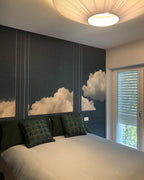 Underside of a Clérachic pleated ceiling light above a bed with dark cushions, in front of a wall mural of clouds.