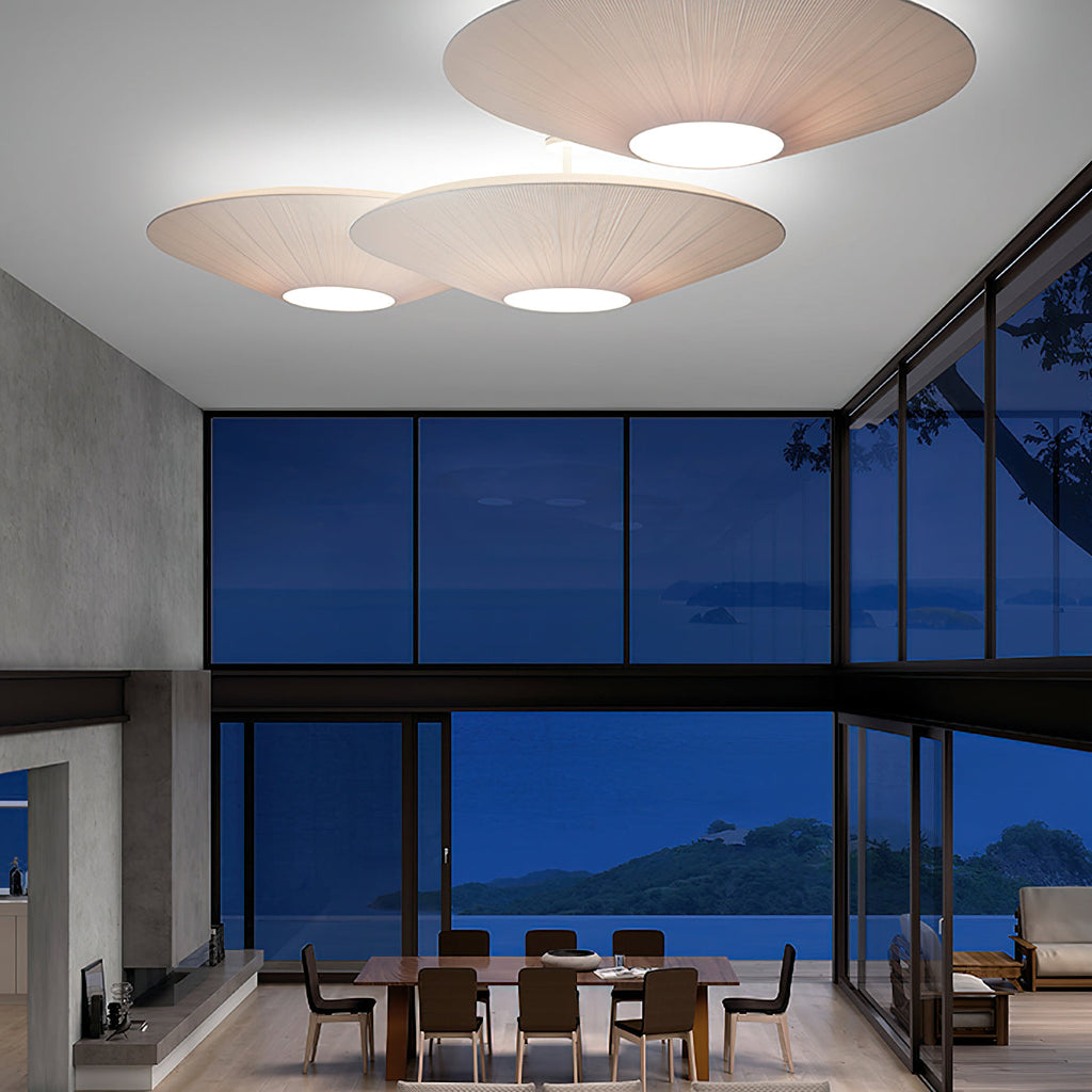 Three Clérachic ivory pleated ceiling lights above a dining table and a corner sofa, with tall glass windows looking over the sea at dusk.
