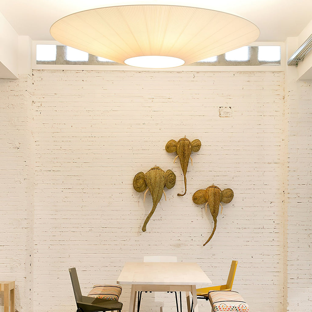 A large Clérachic pleated ceiling light above a white brick wall with three woven animal-head ornaments and a small table below.