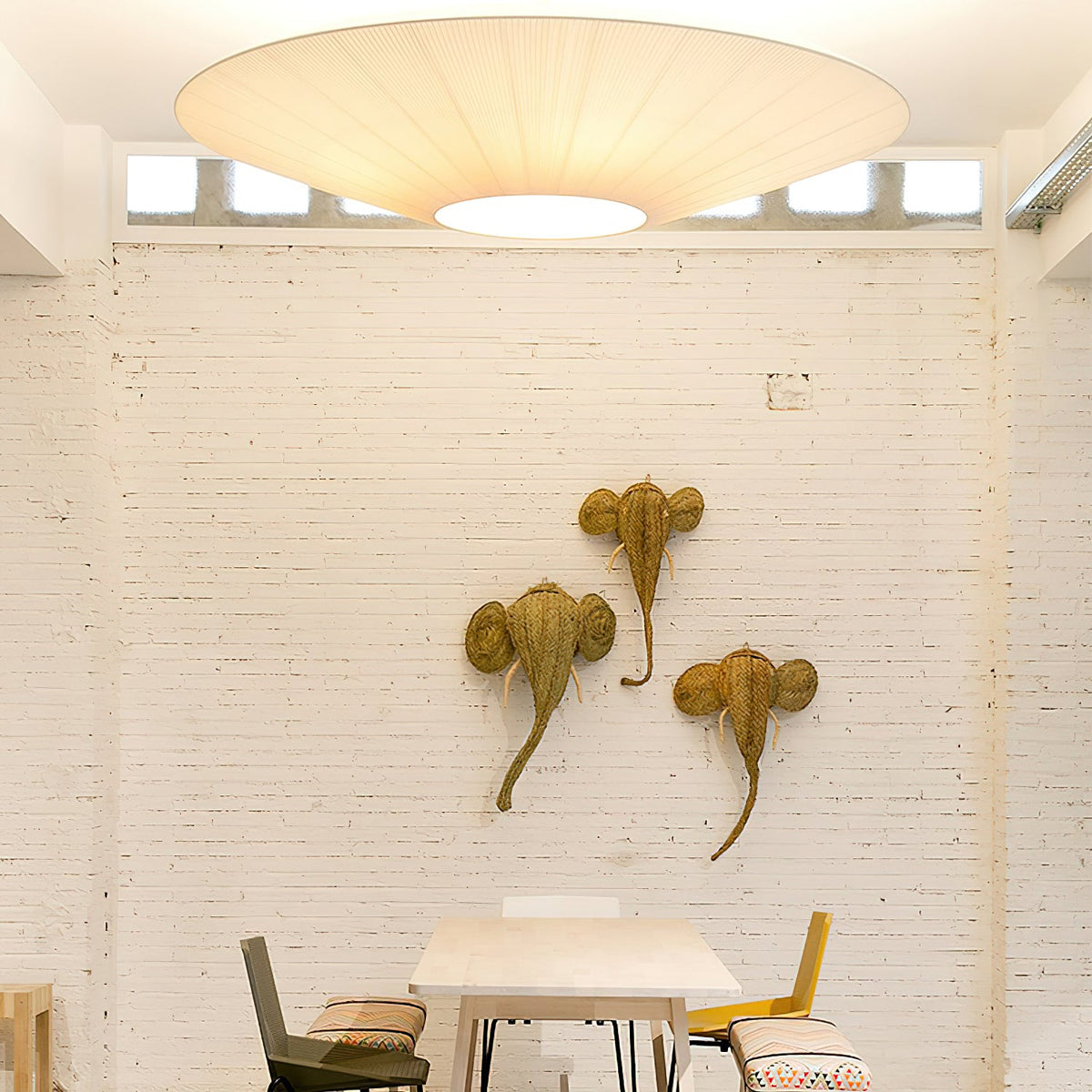 A large Clérachic pleated ceiling light above a white brick wall with three woven animal-head ornaments and a small table below.