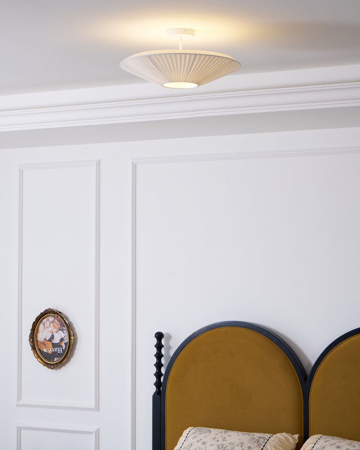 Clérachic pleated ceiling light on a short stem above a white panelled wall, with an arched mustard headboard and an oval framed picture.