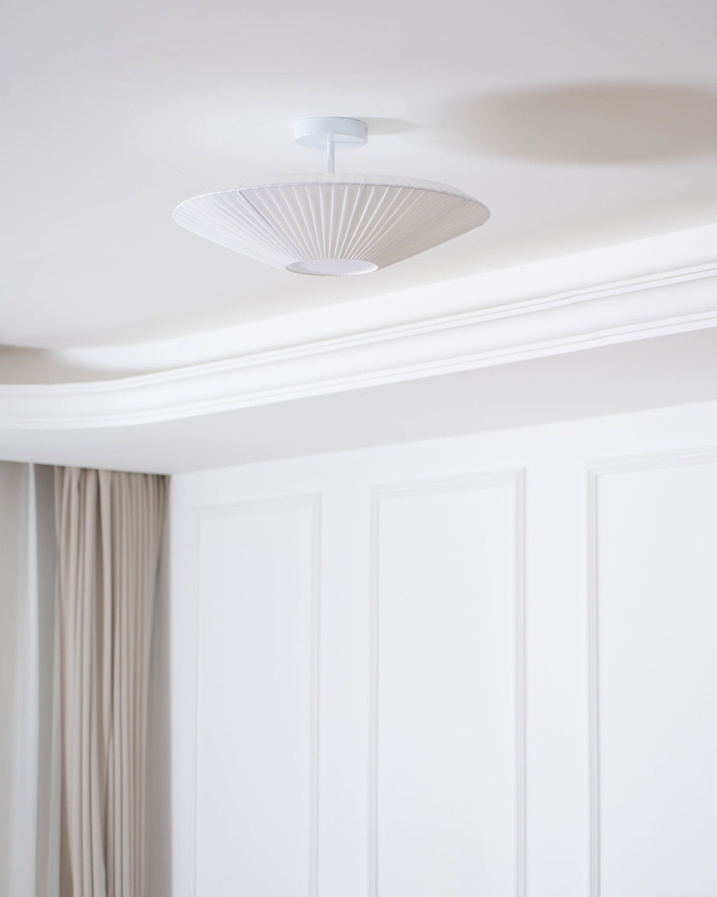 White Clérachic pleated ceiling light on a short stem near a curved cornice, above a panelled wall and long curtains.