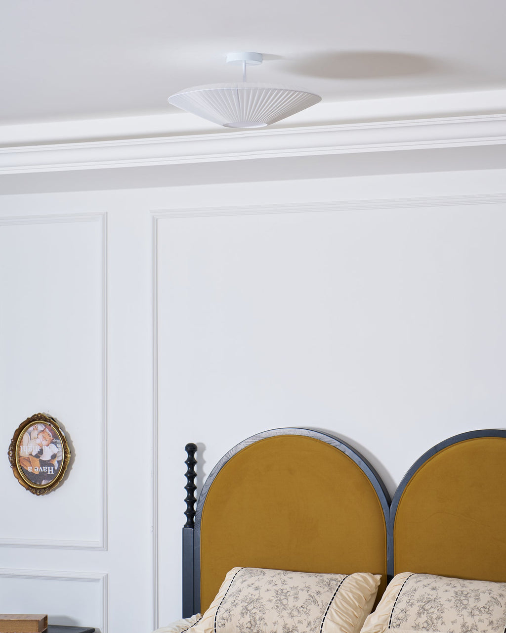 Clérachic pleated ceiling light in white on a short stem, high above a headboard and an oval framed picture on a panelled wall.