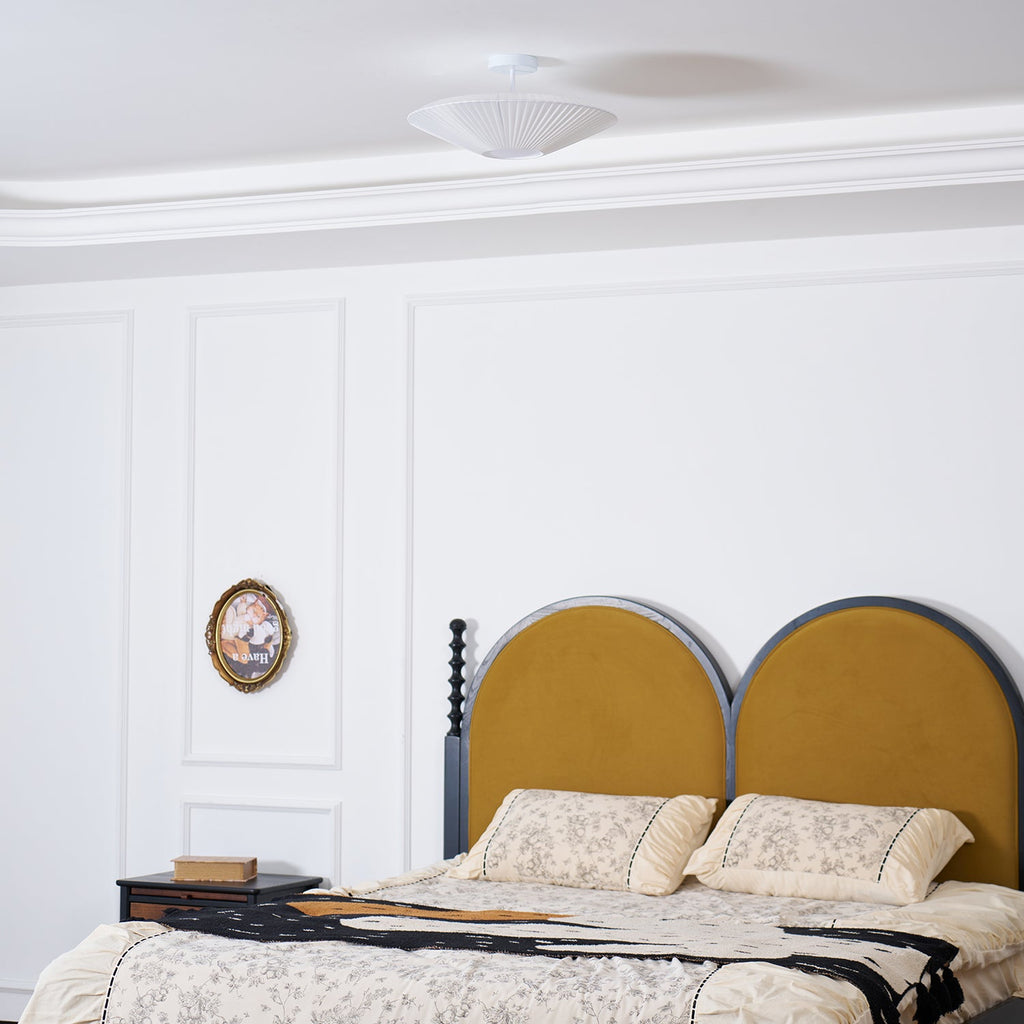 Wider view of a Clérachic white pleated ceiling light above a bed with an arched mustard headboard and floral bedding.
