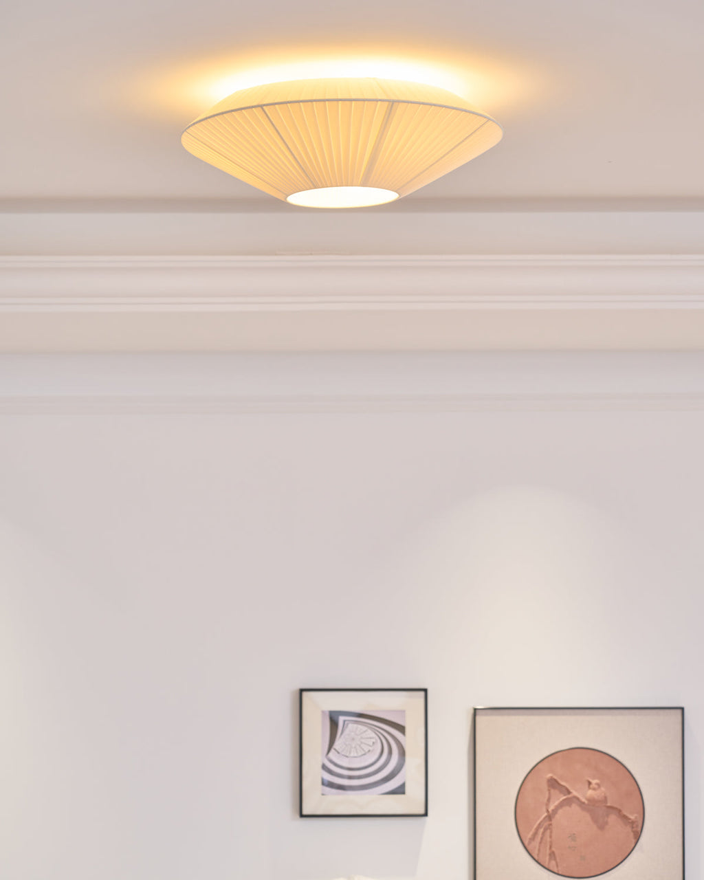Lit Clérachic pleated ceiling light with a warm halo on the ceiling, above a white wall with two framed prints.