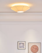 Lit Clérachic pleated ceiling light with a warm halo on the ceiling, above a white wall with two framed prints.