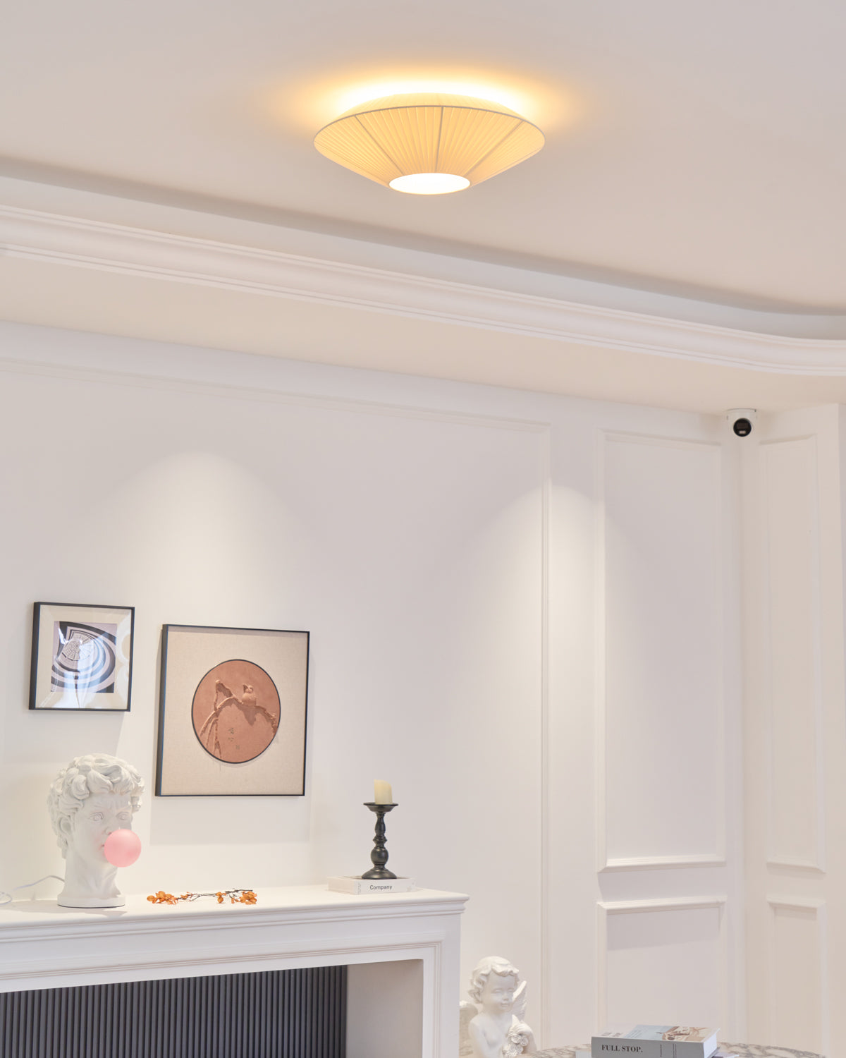 A lit Clérachic pleated ceiling light above a white panelled room with a mantelpiece, two framed prints and a small candlestick.