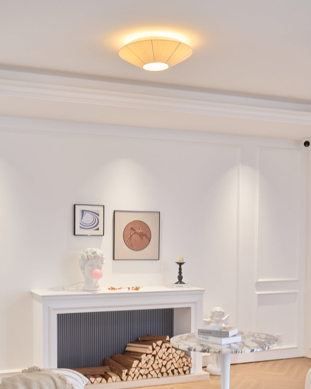 Clérachic pleated ceiling light glowing warmly over a white panelled room with a fireplace, a stack of logs and a small round table.