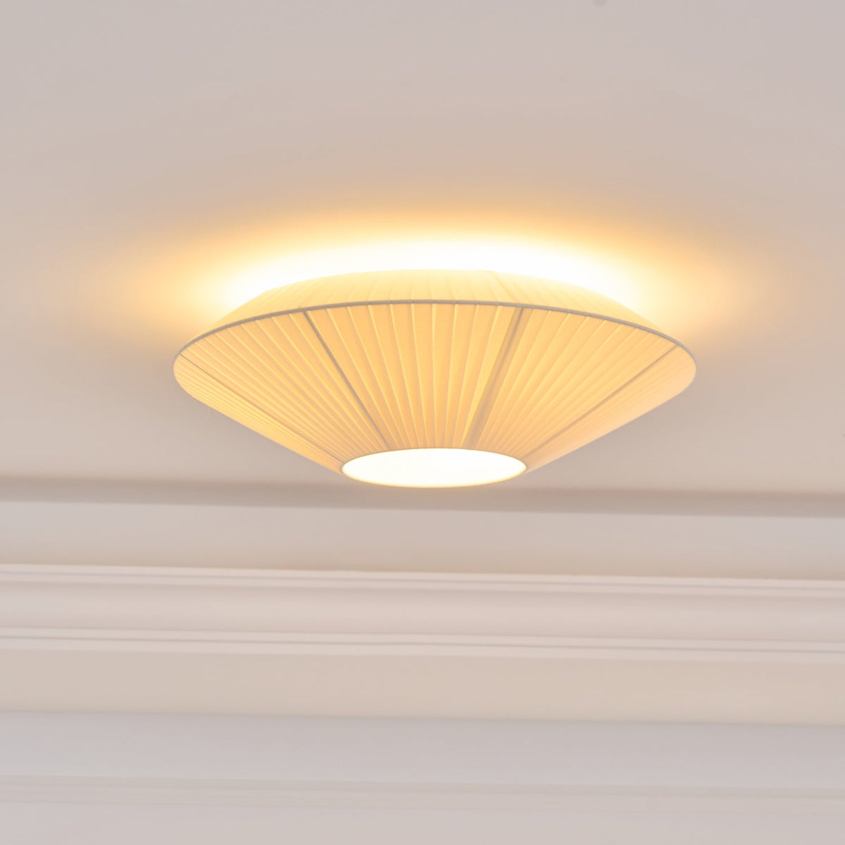 Close view of the lit Clérachic pleated shade, its warm light spilling out around the rim above a moulded cornice.