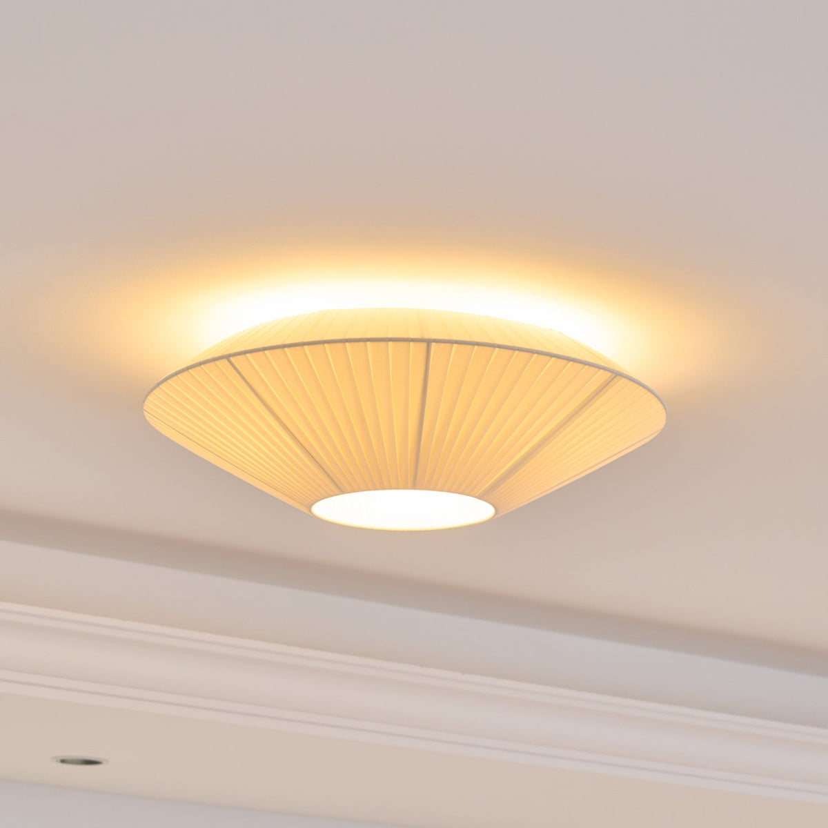 A lit Clérachic pleated ceiling light with seams in its fabric, glowing warm around the rim, above a moulded cornice.