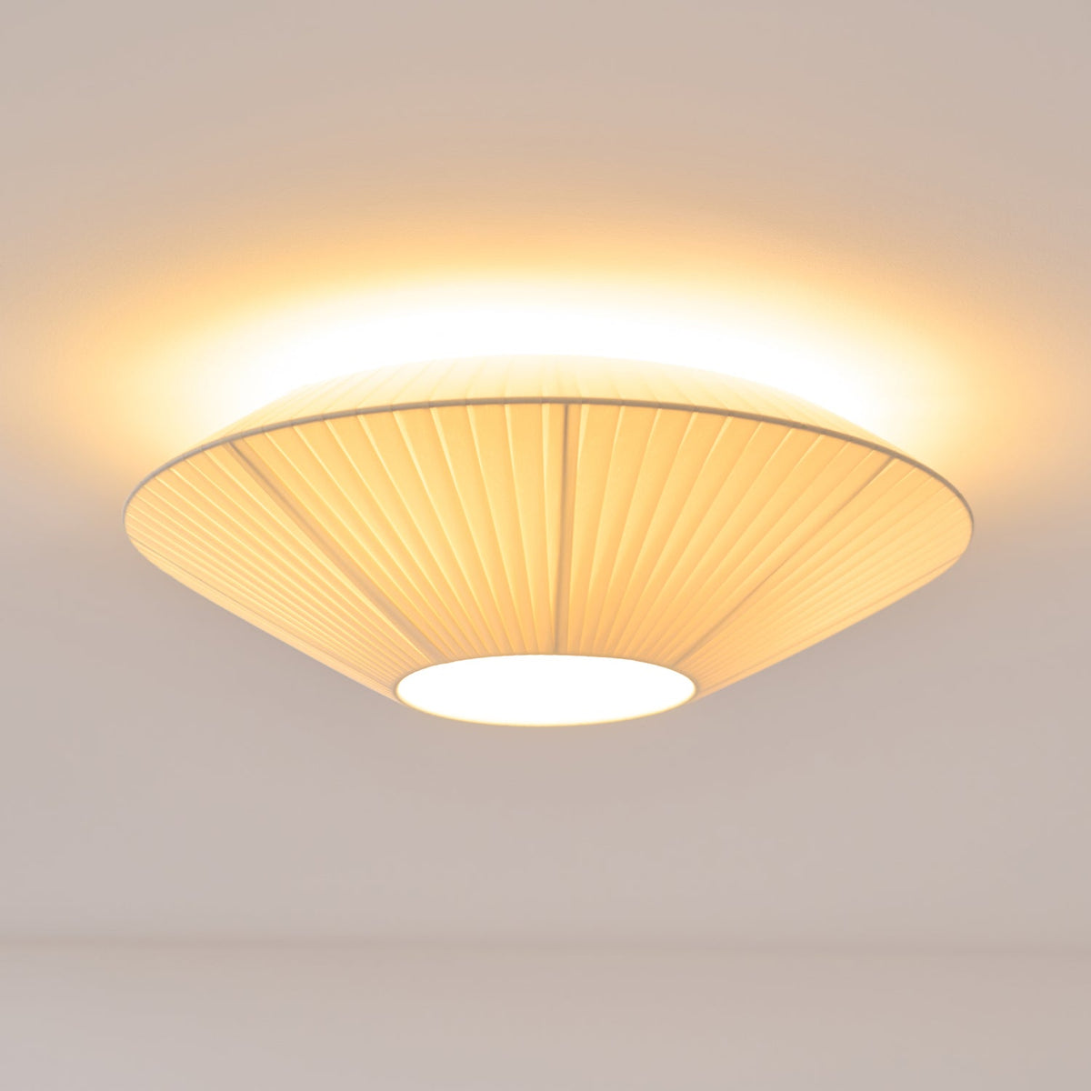 Clérachic pleated ceiling light lit, its pleated shade seen from below with a bright glow around the rim and a white diffuser.