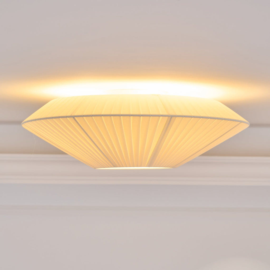 Side view of a lit Clérachic pleated ceiling light, with a wide, flat profile and warm light along the ceiling line.