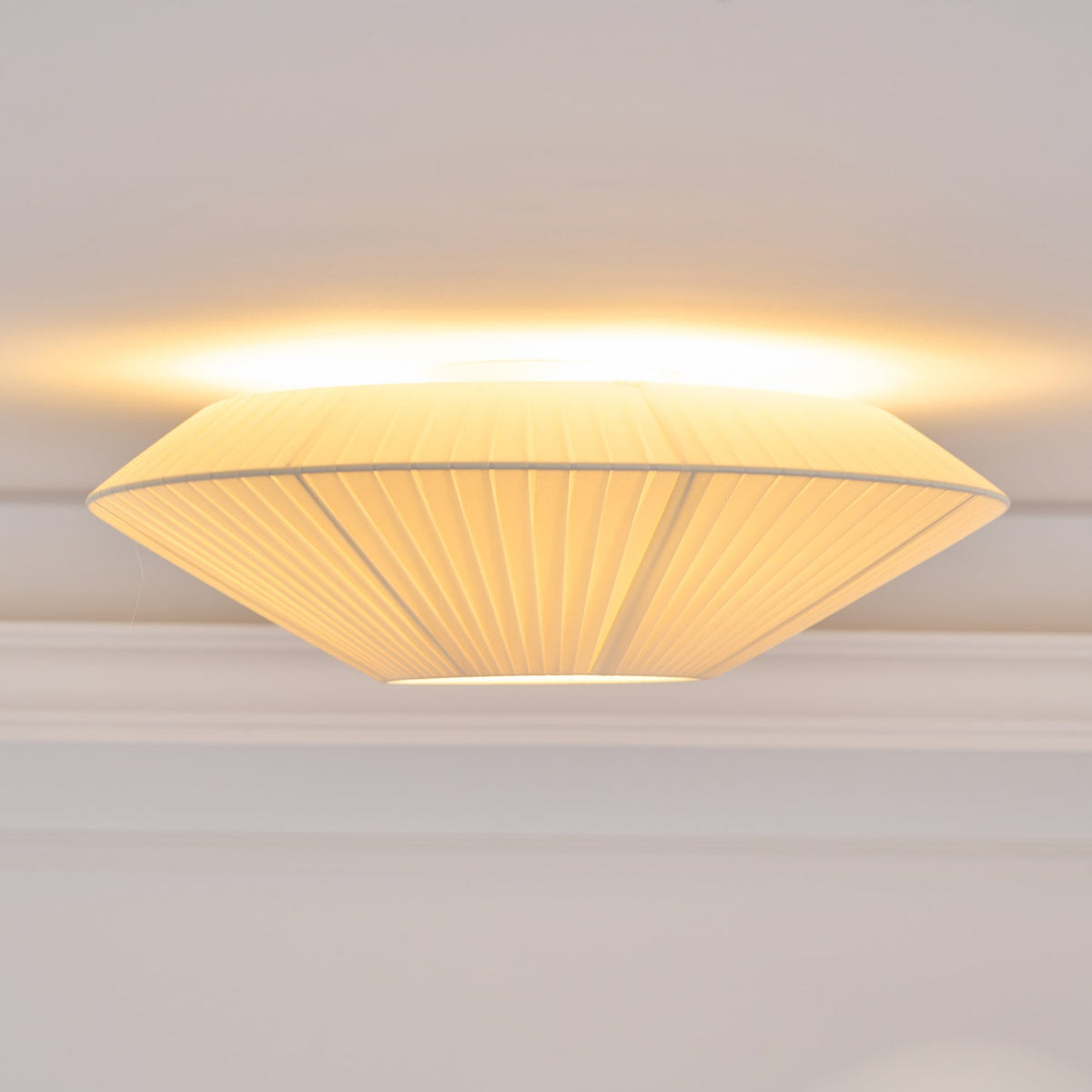Side view of a lit Clérachic pleated ceiling light, with a wide, flat profile and warm light along the ceiling line.