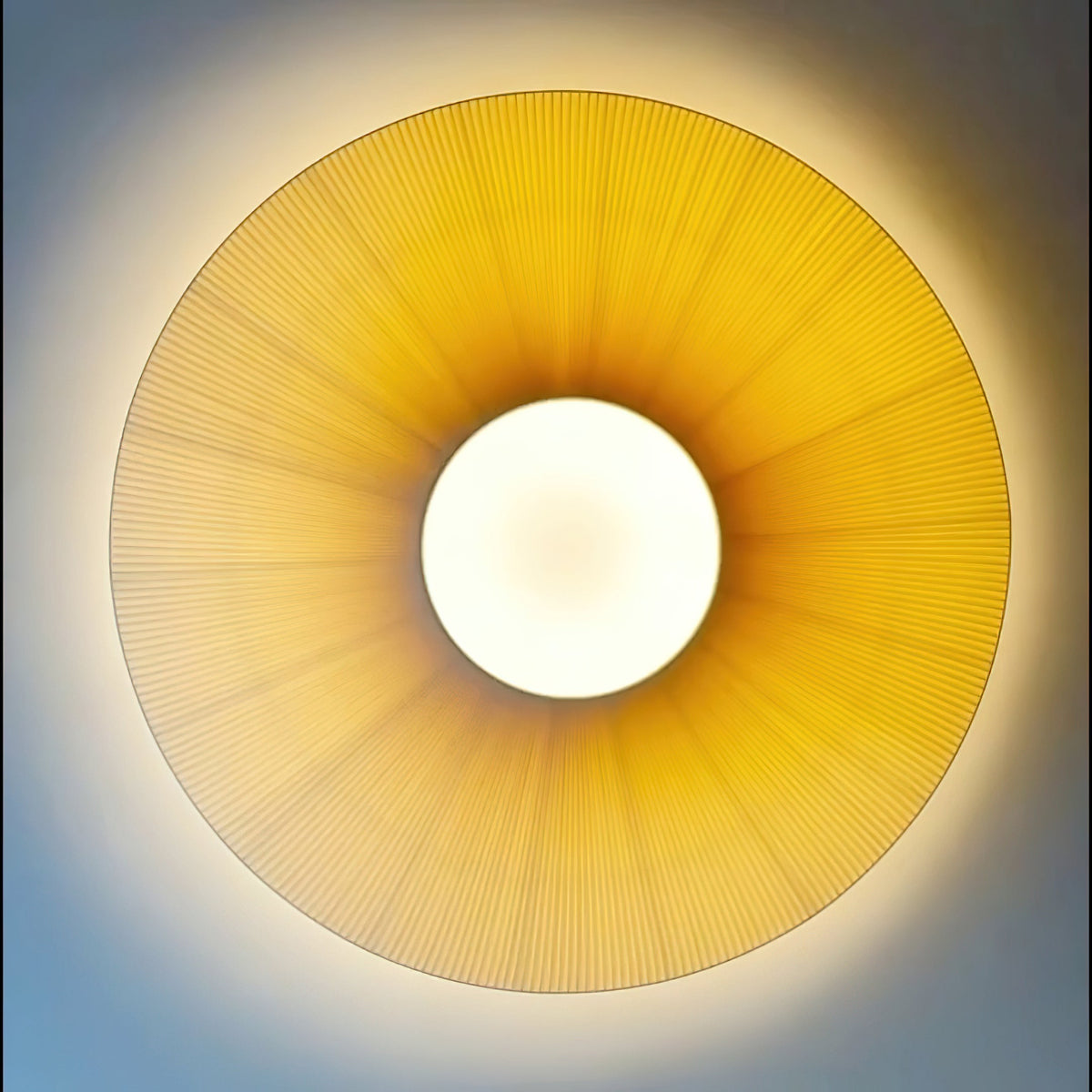 Looking straight up at a lit Clérachic pleated ceiling light, a golden ring of fabric around a white round diffuser.