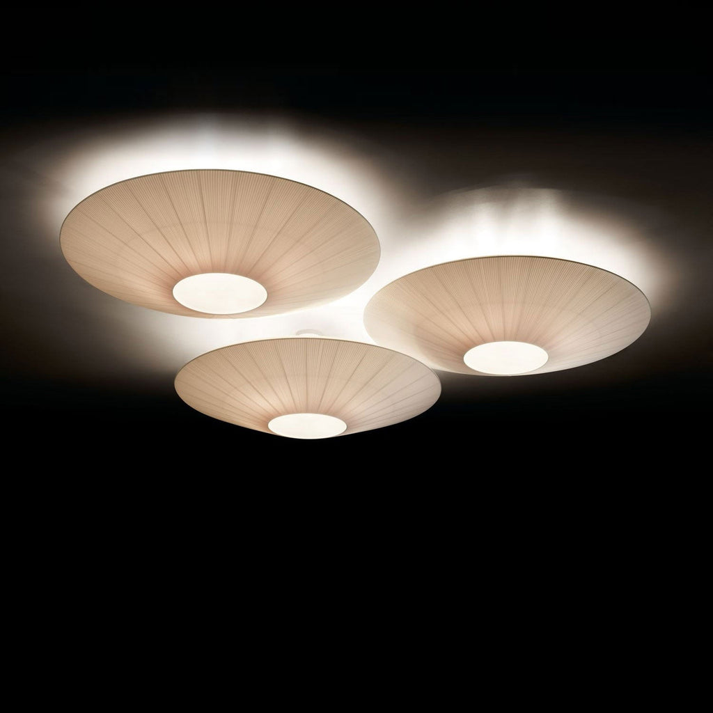 Three Clérachic pleated ceiling lights in ivory, lit against a black background, with white light glowing around their rims.