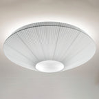 Lit Clérachic pleated ceiling light in grey and white, with a soft glow around the rim against a pale wall.