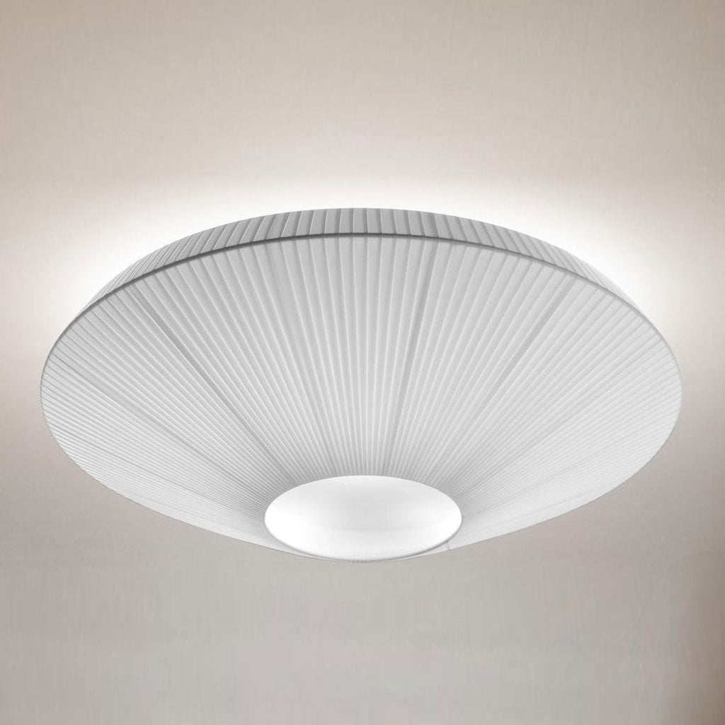 A Clérachic pleated ceiling light in white with a grey pleated edge and a white diffuser, lit softly against a taupe wall.