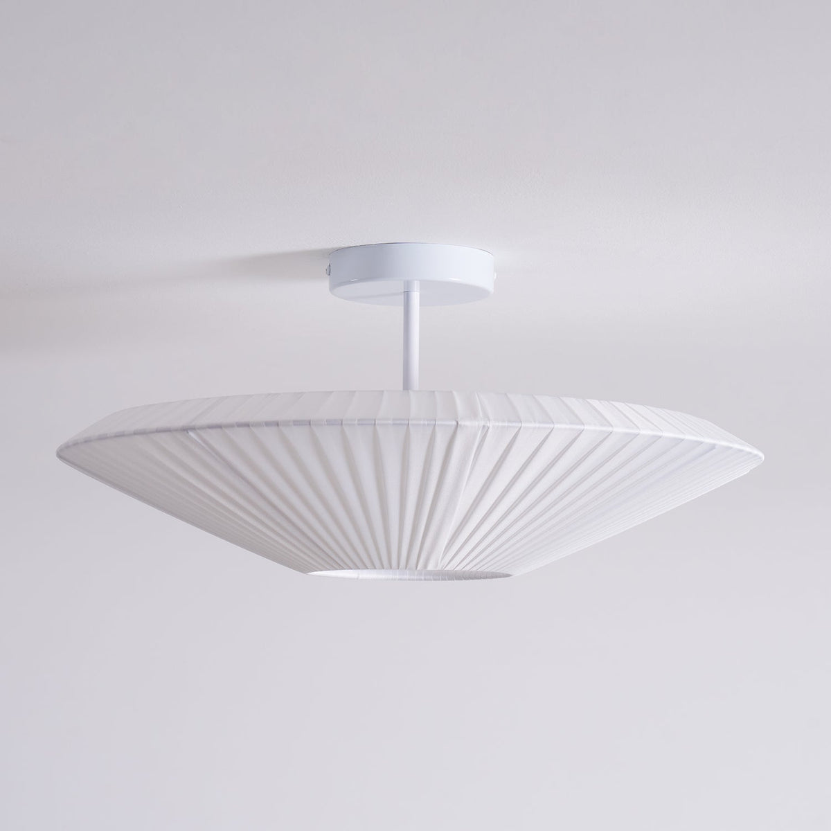 Side view of an unlit Clérachic white pleated ceiling light on a straight stem, with a flat, wide profile against a pale grey wall.