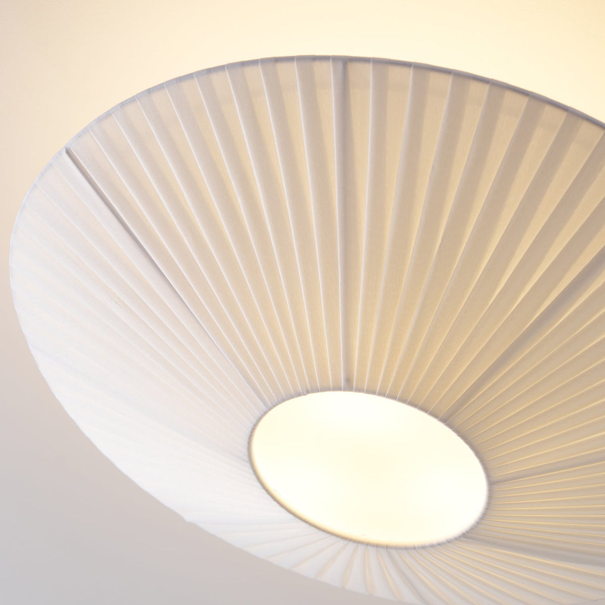 Close view from below of the pleated white fabric and round diffuser on this Clérachic ceiling light, lit warmly.