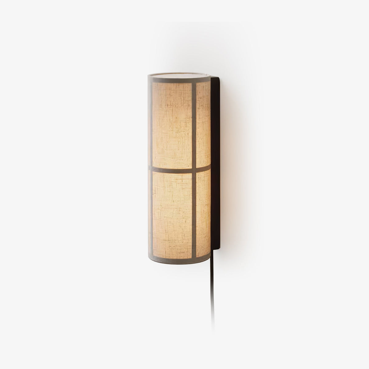 This Clérachic plug-in wall light on a white backdrop, a slim fabric cylinder shade glowing warm in a black frame, with its cord hanging below.