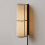 Close view of a lit Clérachic fabric cylinder wall light on a pale grey wall, its linen shade crossed by two bands and a black back plate.