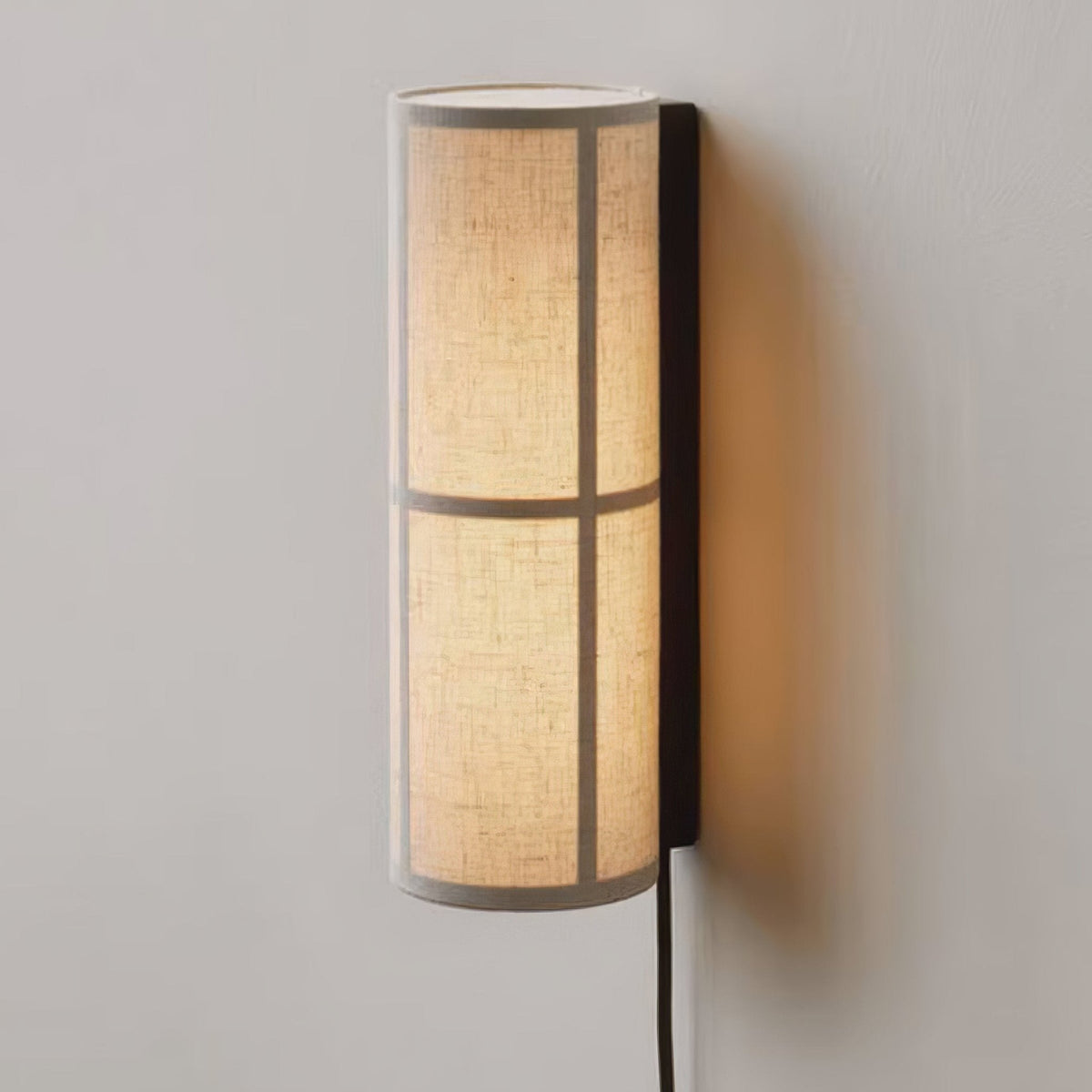 Close view of a lit Clérachic fabric cylinder wall light on a pale grey wall, its linen shade crossed by two bands and a black back plate.