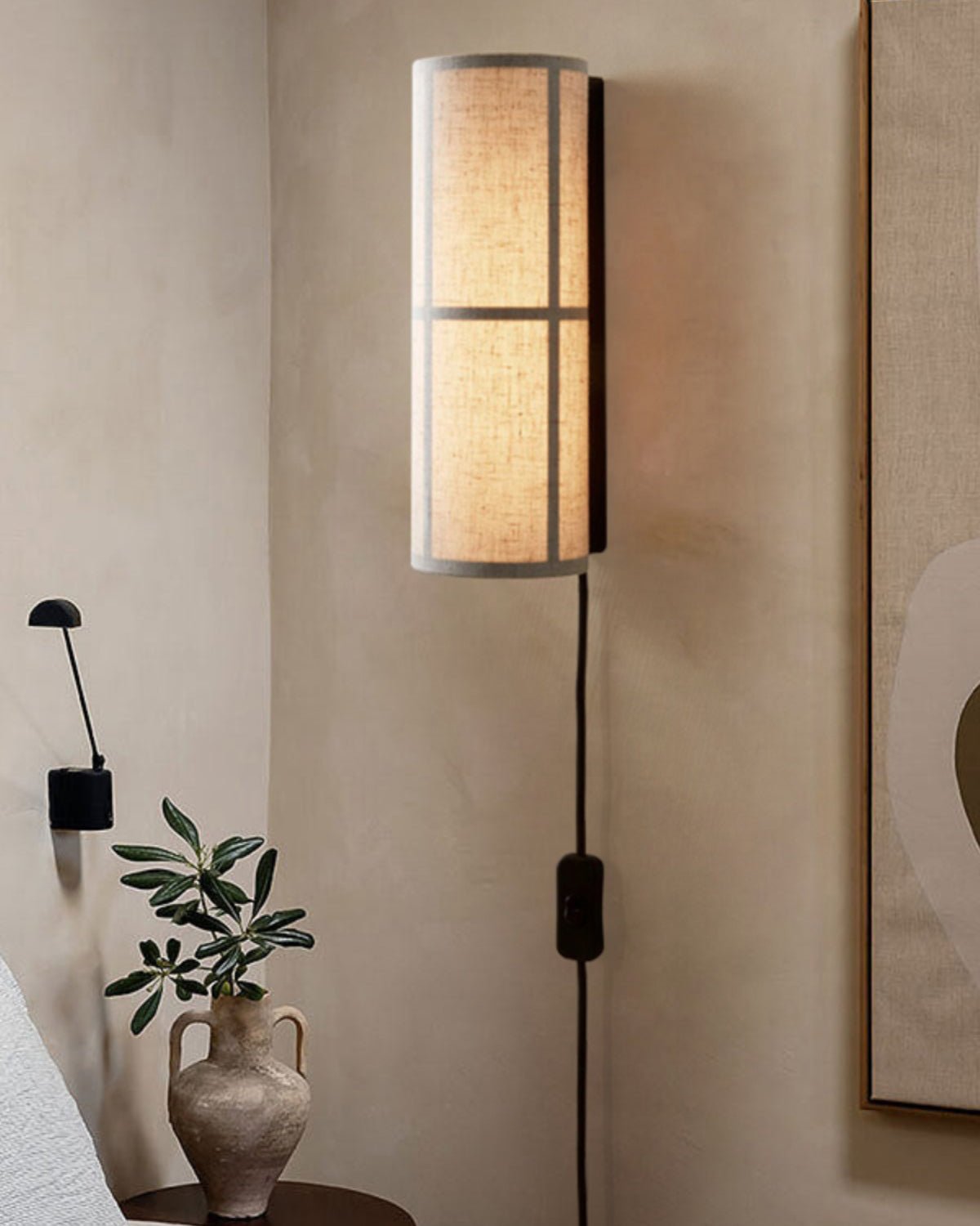 Clérachic fabric wall light lit above a bedside table with a clay vase and olive stem, its black cord and inline switch hanging down.