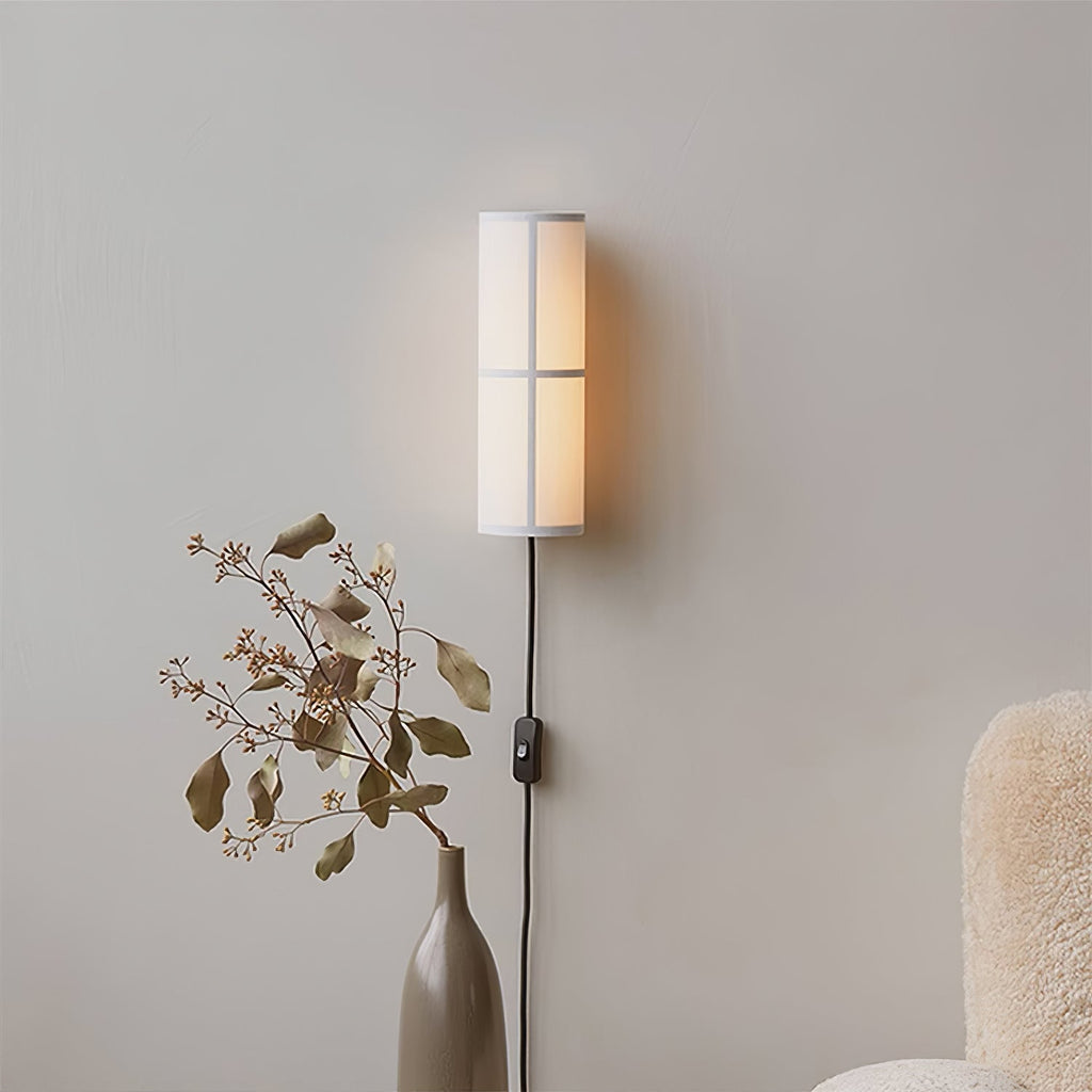 A white-shaded Clérachic cylinder wall light glows softly on a grey wall, above a tall vase of dried stems and a fluffy chair.