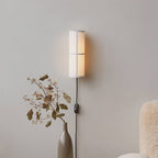 A white-shaded Clérachic cylinder wall light glows softly on a grey wall, above a tall vase of dried stems and a fluffy chair.