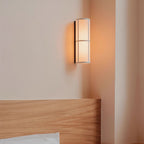 Orange evening light from a Clérachic fabric cylinder wall light on a peach wall, just above a pale wood headboard.