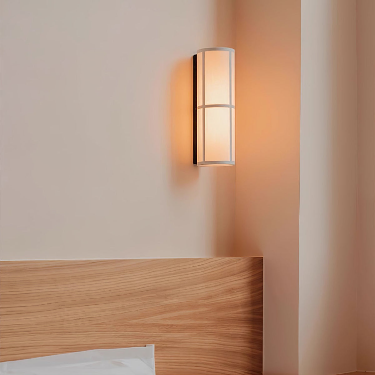 Orange evening light from a Clérachic fabric cylinder wall light on a peach wall, just above a pale wood headboard.