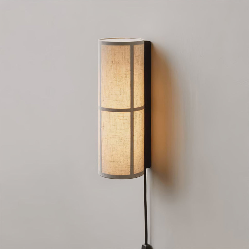 Slim Clérachic cylinder wall light on a pale grey wall, its linen shade glowing from within and a black cord running down.