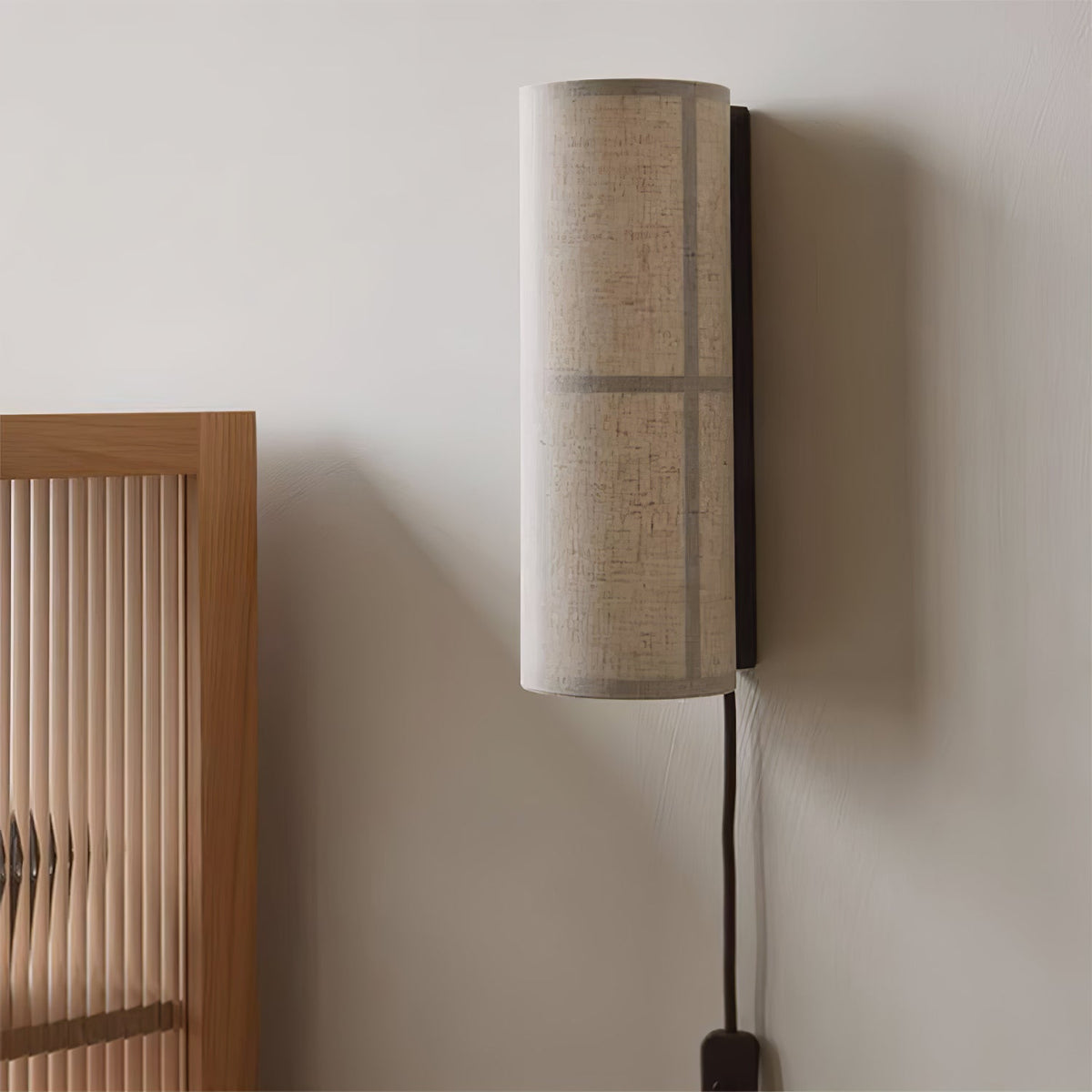 Side view of this Clérachic wall light on a grey wall beside a fluted wooden cabinet, its linen cylinder shade standing out from the black back plate.