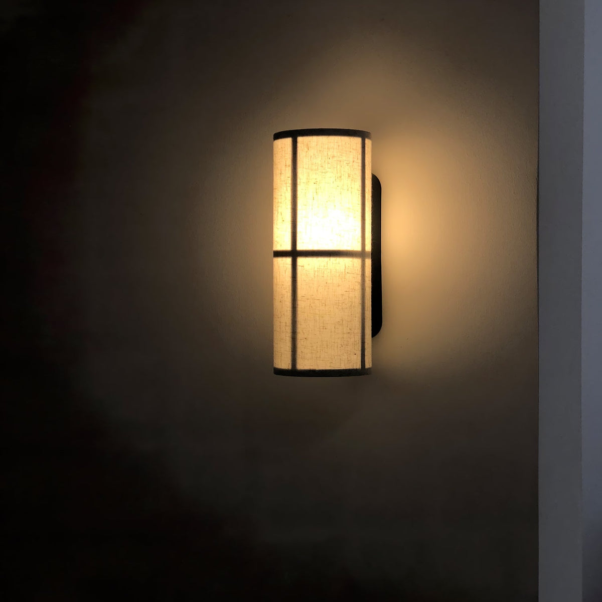 A Clérachic cylinder wall light lit on a dark wall, its linen shade glowing amber with a soft halo around the black frame.