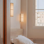 Lit Clérachic wall light on a bedroom wall by a window, with a second light beyond a doorway and a white pillow below.