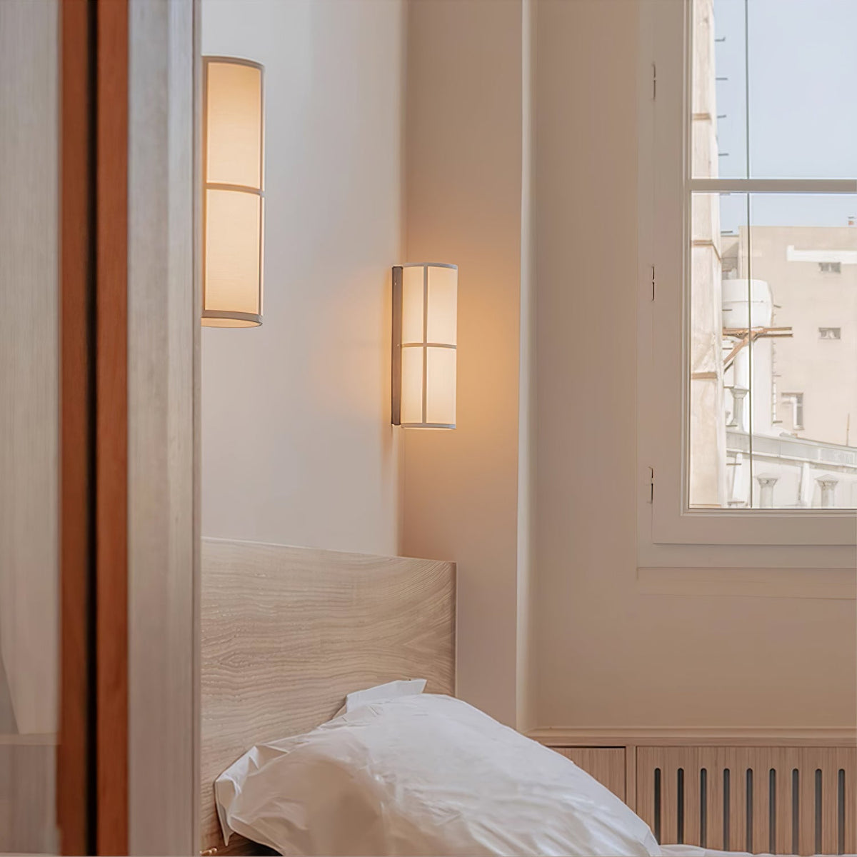 Lit Clérachic wall light on a bedroom wall by a window, with a second light beyond a doorway and a white pillow below.