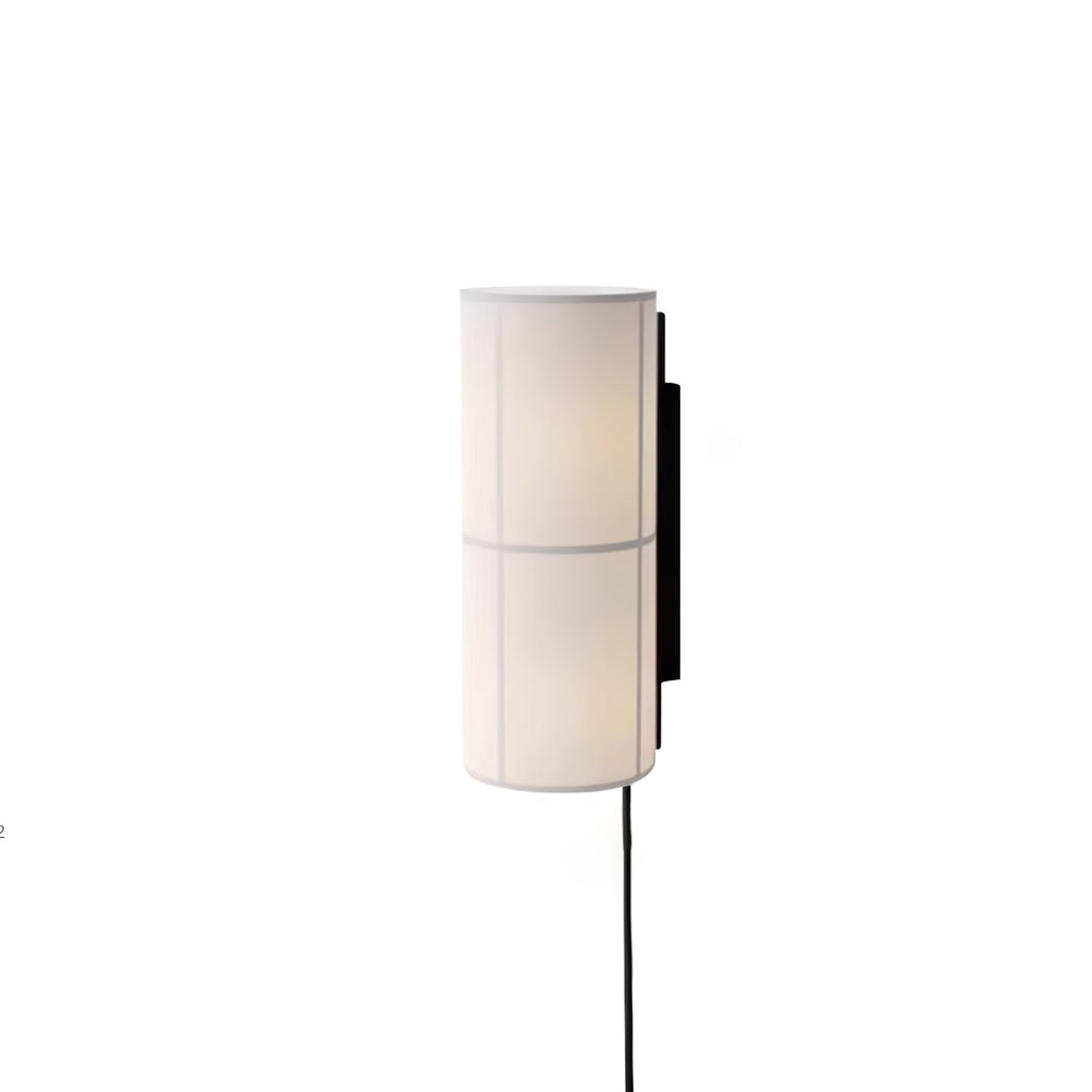 A white Clérachic cylinder wall light, softly lit, on a white backdrop with a black cord hanging below it.