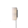 A white Clérachic cylinder wall light, softly lit, on a white backdrop with a black cord hanging below it.