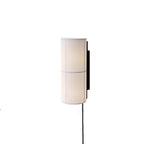 A white Clérachic cylinder wall light, softly lit, on a white backdrop with a black cord hanging below it.