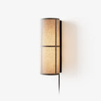 Clérachic fabric wall light lit against a pale backdrop, its linen shade glowing warm and its black cord trailing below.