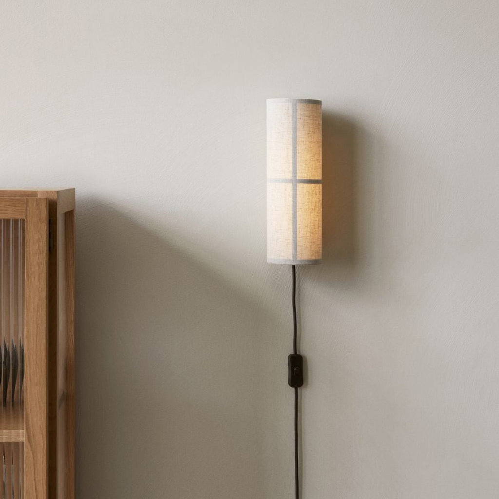 A grey linen Clérachic cylinder wall light on a pale wall beside a wooden cabinet, its black cord and inline switch hanging below.