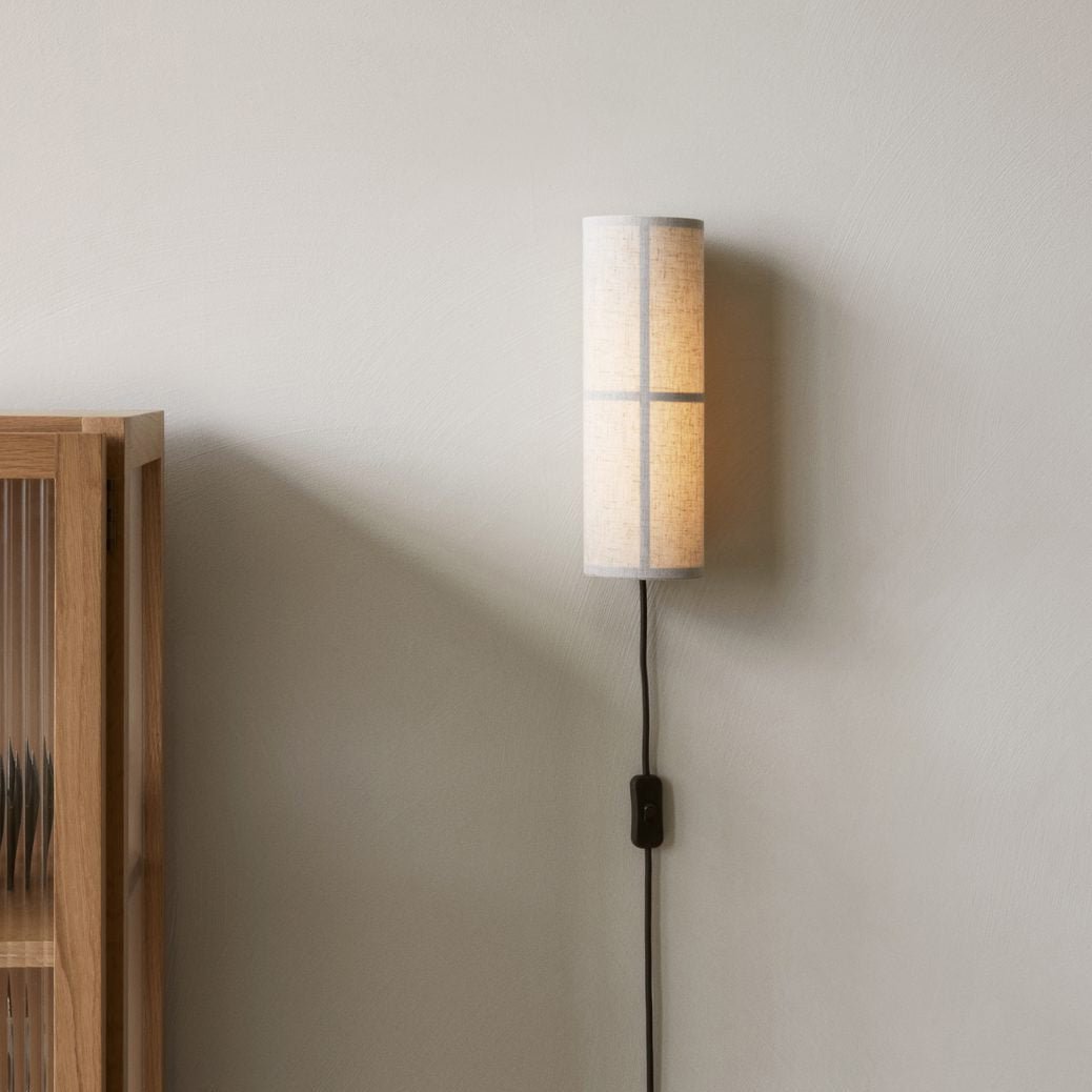 A grey linen Clérachic cylinder wall light on a pale wall beside a wooden cabinet, its black cord and inline switch hanging below.