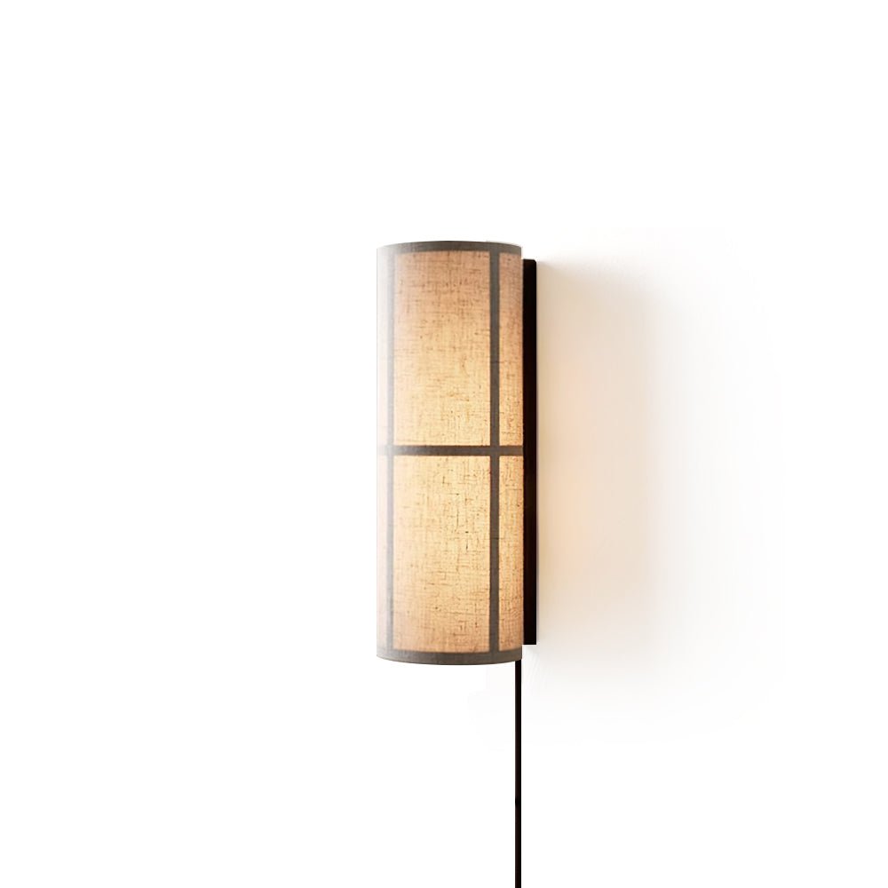 Lit Clérachic cylinder wall light with a warm glow behind the linen shade, its black cord falling straight down on white.