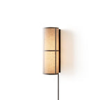 Lit Clérachic cylinder wall light with a warm glow behind the linen shade, its black cord falling straight down on white.