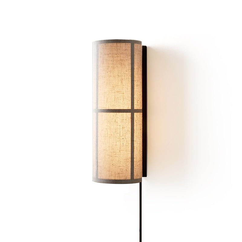Large view of a lit Clérachic cylinder wall light, the banded linen shade glowing in a black frame, with a cord hanging below.